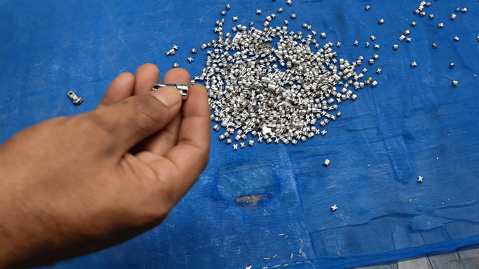 Hand holding a shiny silver nickel-plated small hardware component with scattered similar parts on a blue background.